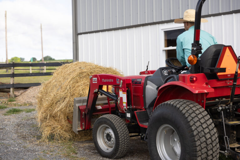 A farmer driving a red Mahindra tractor is transporting a large round bale of hay. The bale is secured on the front loader attachment as the farmer approaches a metal barn. The scene highlights the efficient use of hay transport equipment in farm operations. The background shows a fenced pasture, and a horse stands nearby observing. The overcast sky and the farmer's straw hat give the impression of a routine day on the farm, focused on preparing feed for the livestock.