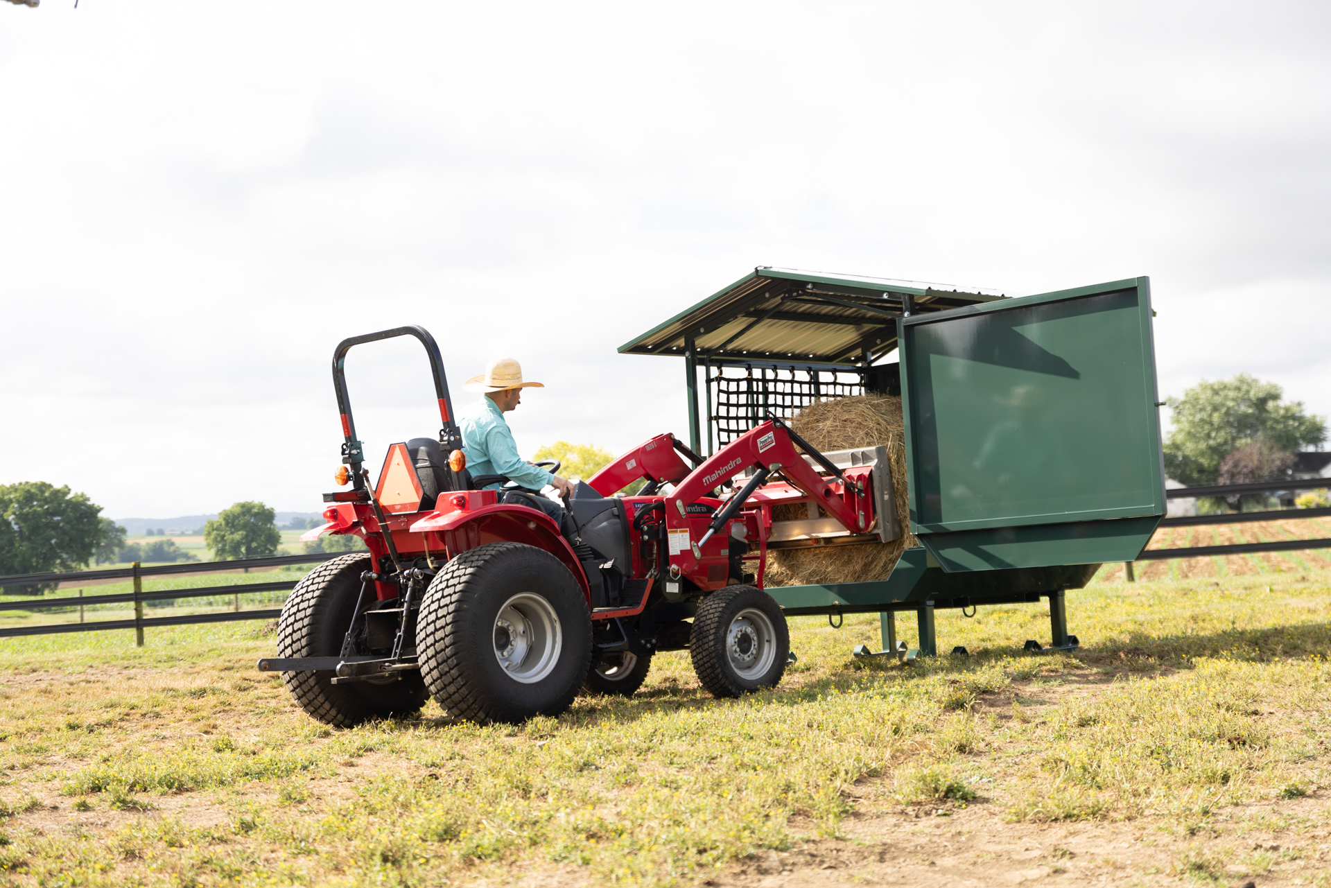 A farmer, wearing a cowboy-style hat, operates a red Mahindra tractor equipped with a front loader attachment to transport a large hay bale. The tractor is positioned next to a green hay feeder, where the bale is unloaded. The scene occurs on a grassy field with a wooden fence and a scenic rural backdrop of trees and hills.