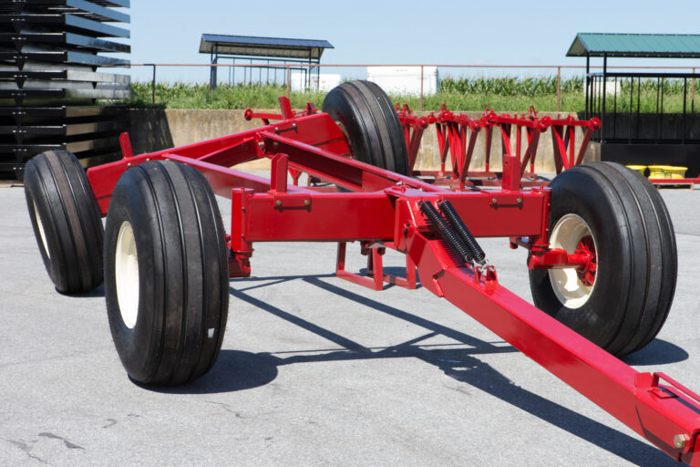 A close-up view of a red hay transport trailer, designed for hauling large bales of hay. The equipment features sturdy wheels and a solid frame, showcasing its durability and ability to handle heavy loads. The trailer is parked on a concrete surface under a clear blue sky, with agricultural structures and greenery in the background, emphasizing its purpose on a farm. This trailer is an essential piece of equipment for efficiently transporting hay across fields or farmyards.